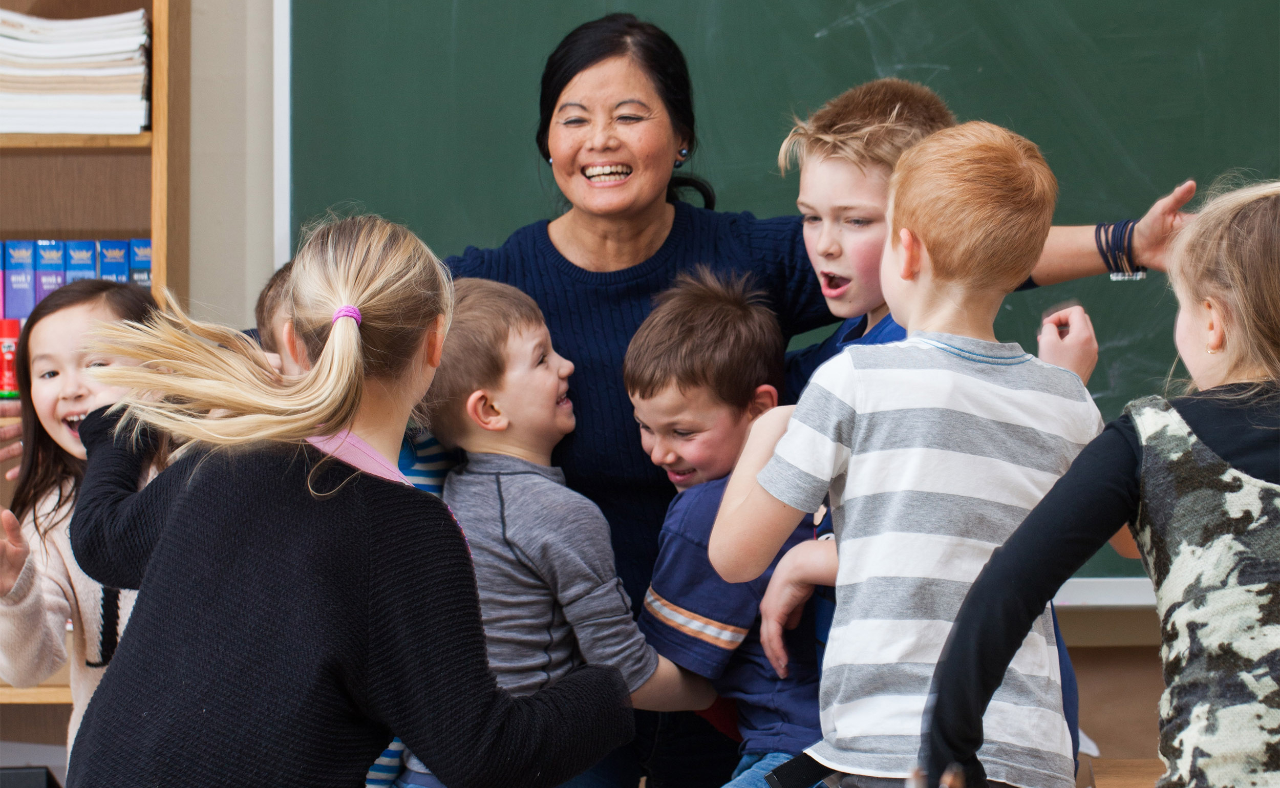 Lærer omringet av glade barn foran en grønn tavle i klasserommet